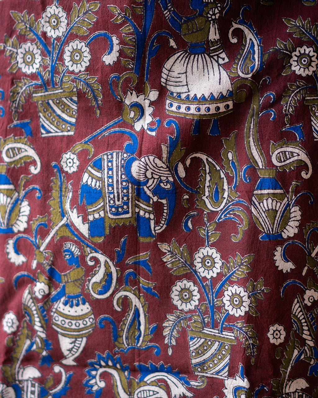 A close-up of a patterned textile featuring a traditional Indian block print design with a blue elephant, white flowers, and ornate paisley motifs on a deep maroon background.