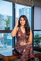Woman in a patterned dress sitting by a window with a cityscape view, holding a mug.