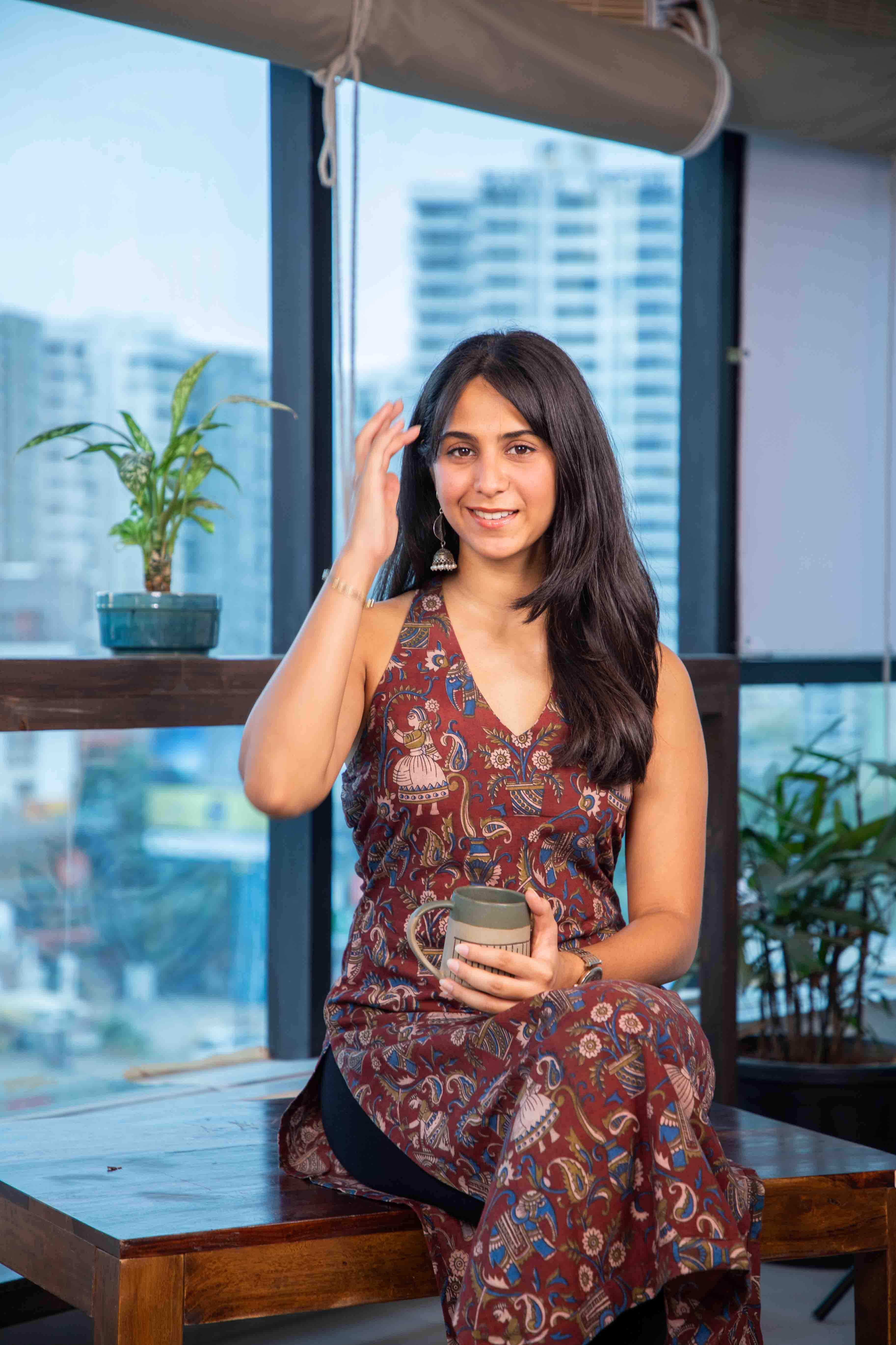 Woman in a patterned dress sitting by a window with a cityscape view, holding a mug.