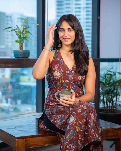 Woman in a patterned dress sitting by a window with a cityscape view, holding a mug.