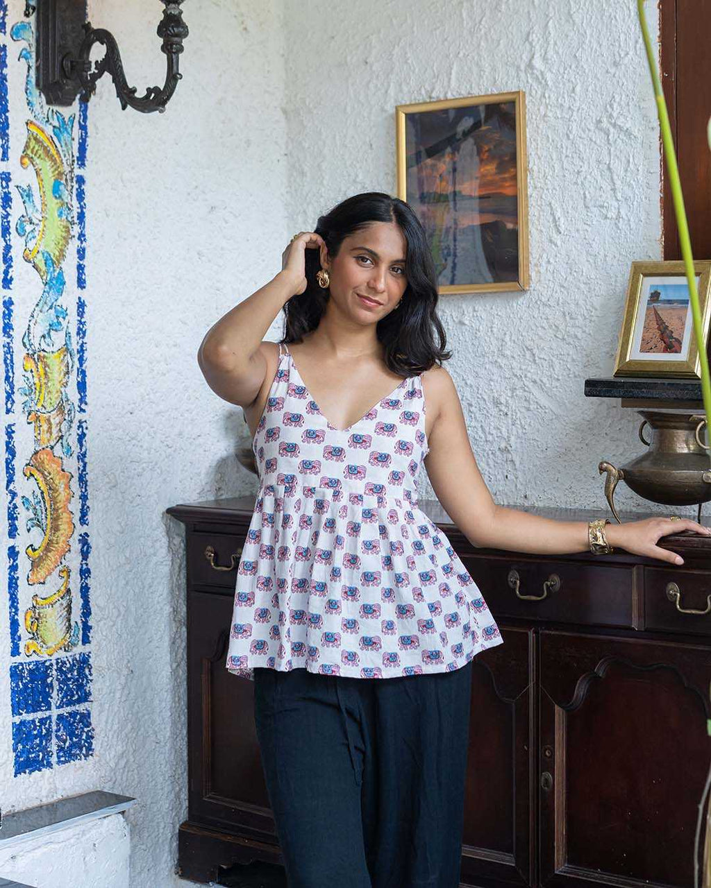 Woman in a patterned top and dark pants standing in a room with a white wall and wooden furniture.